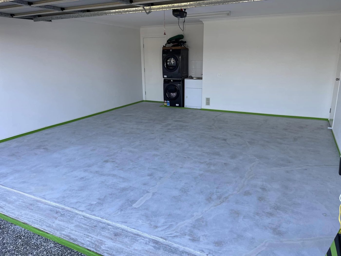 Garage floor before coating, bare grey concrete with green masking tape running along the base of the white walls and a stacked washer and dryer in the back corner.