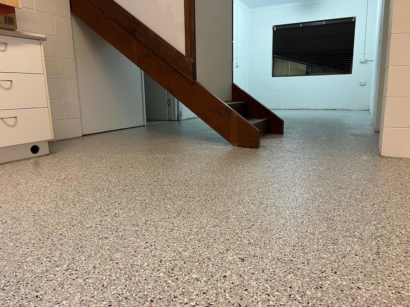 Flood affected downstairs room after coating, an even beige and black flake floor running seamlessly under the stairwell, past the cabinets and through the doorway into the next room.