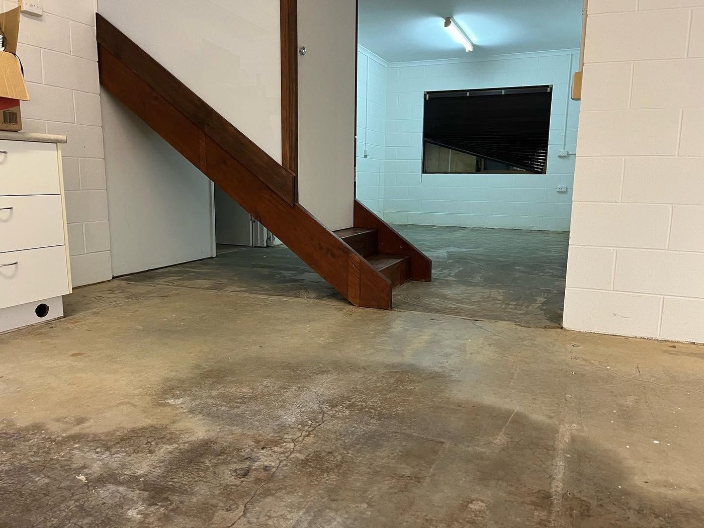 The same downstairs room before coating, a stained and patchy concrete slab running under a timber stairwell and through to a second room with painted block walls.