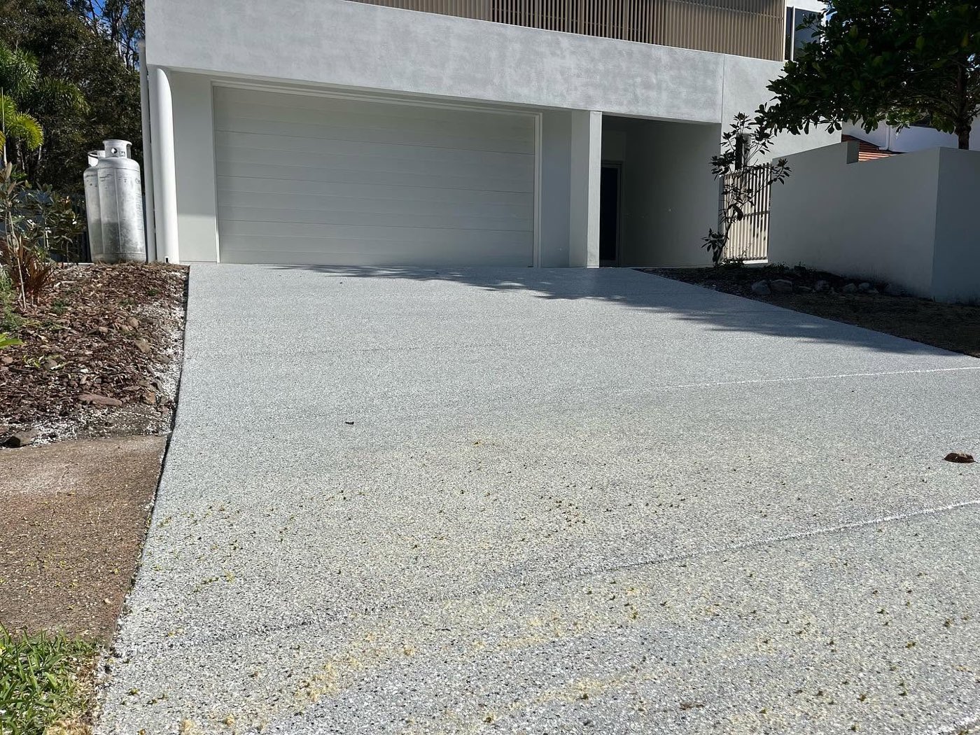 Finished residential driveway coated in a pale grey speckled non slip finish, running in full sun up to a closed white garage door.