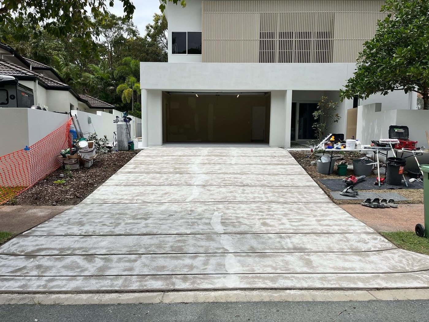 The same driveway before coating, ground back to raw concrete with white grinding residue across the slab, prep gear and buckets set up on the lawn beside it.