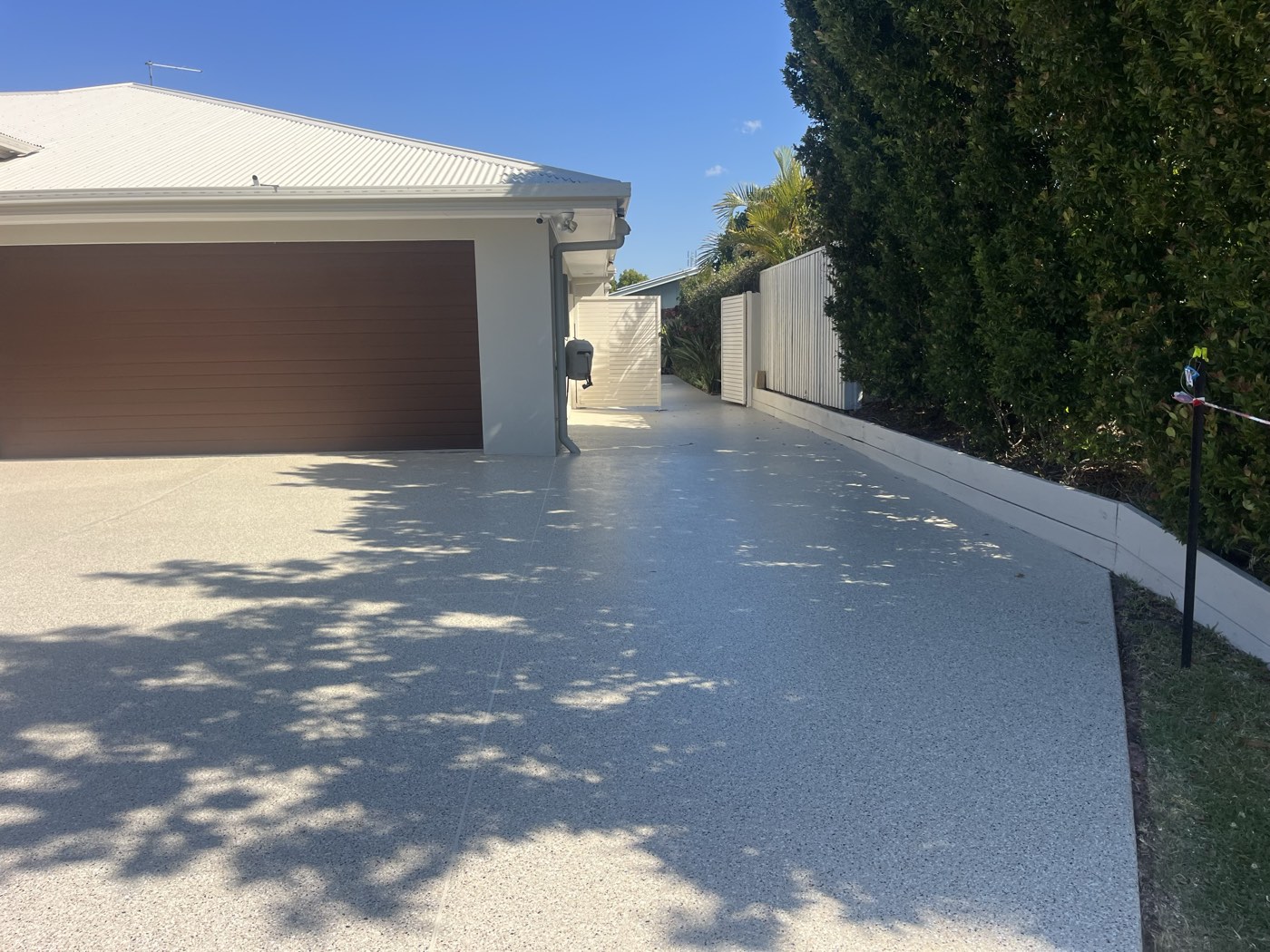 The same driveway after coating, a light grey and white flake finish running from the brown garage door down the side of the house beside a tall hedge.