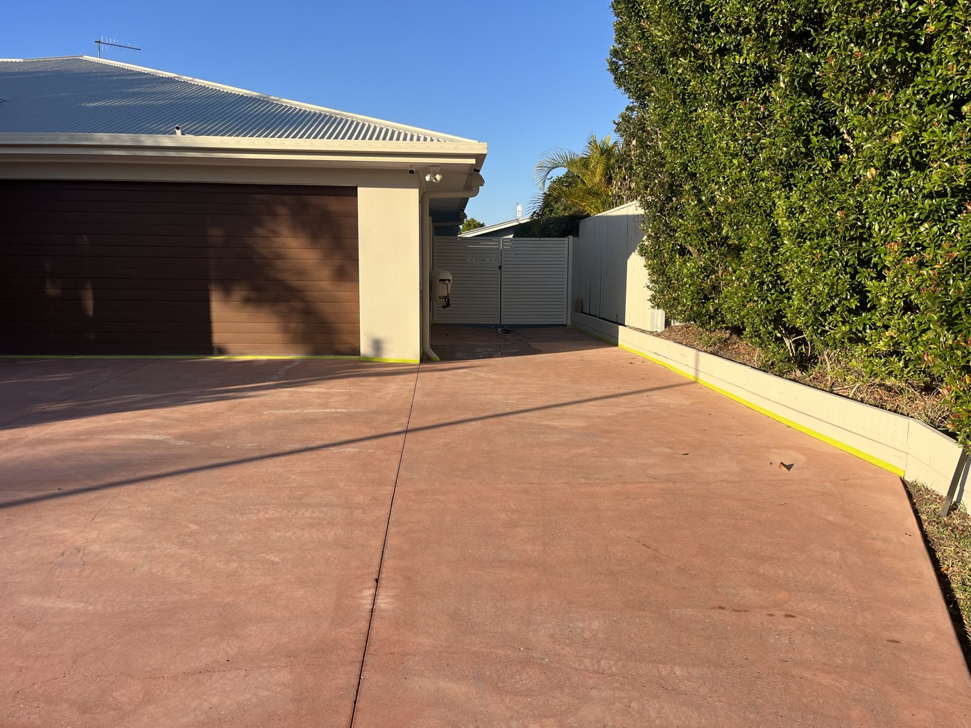 Driveway before coating, faded red coloured concrete in front of a double garage with a brown panel door and a tall hedge along the right hand side.