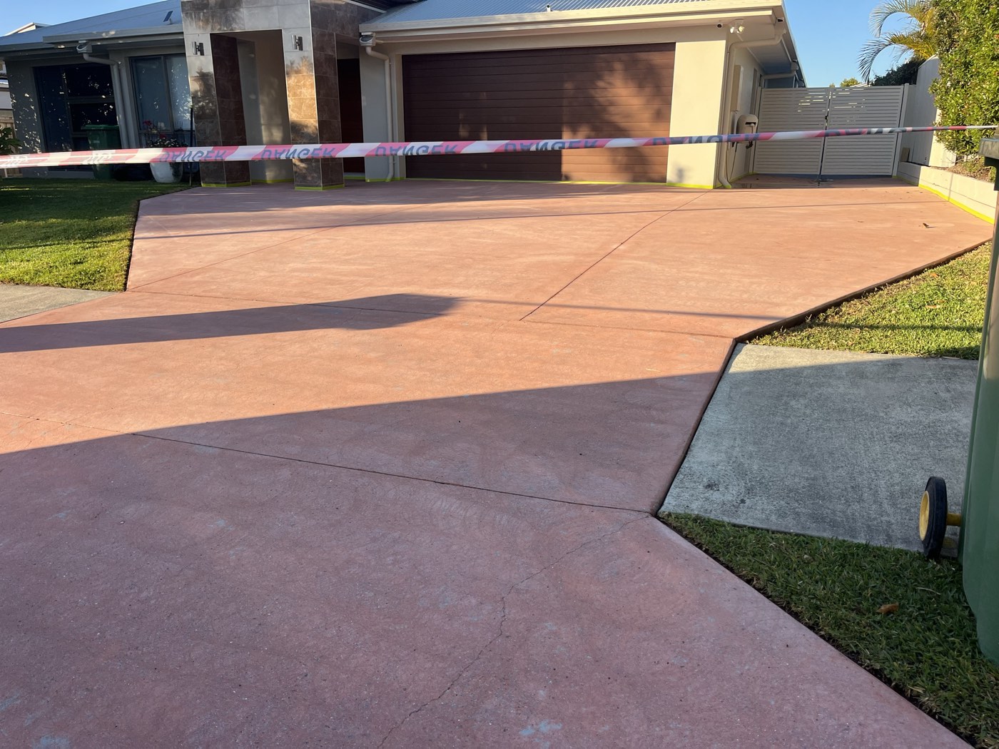 Driveway before coating, faded red coloured concrete taped off with barrier tape in front of a double garage with stone clad pillars.