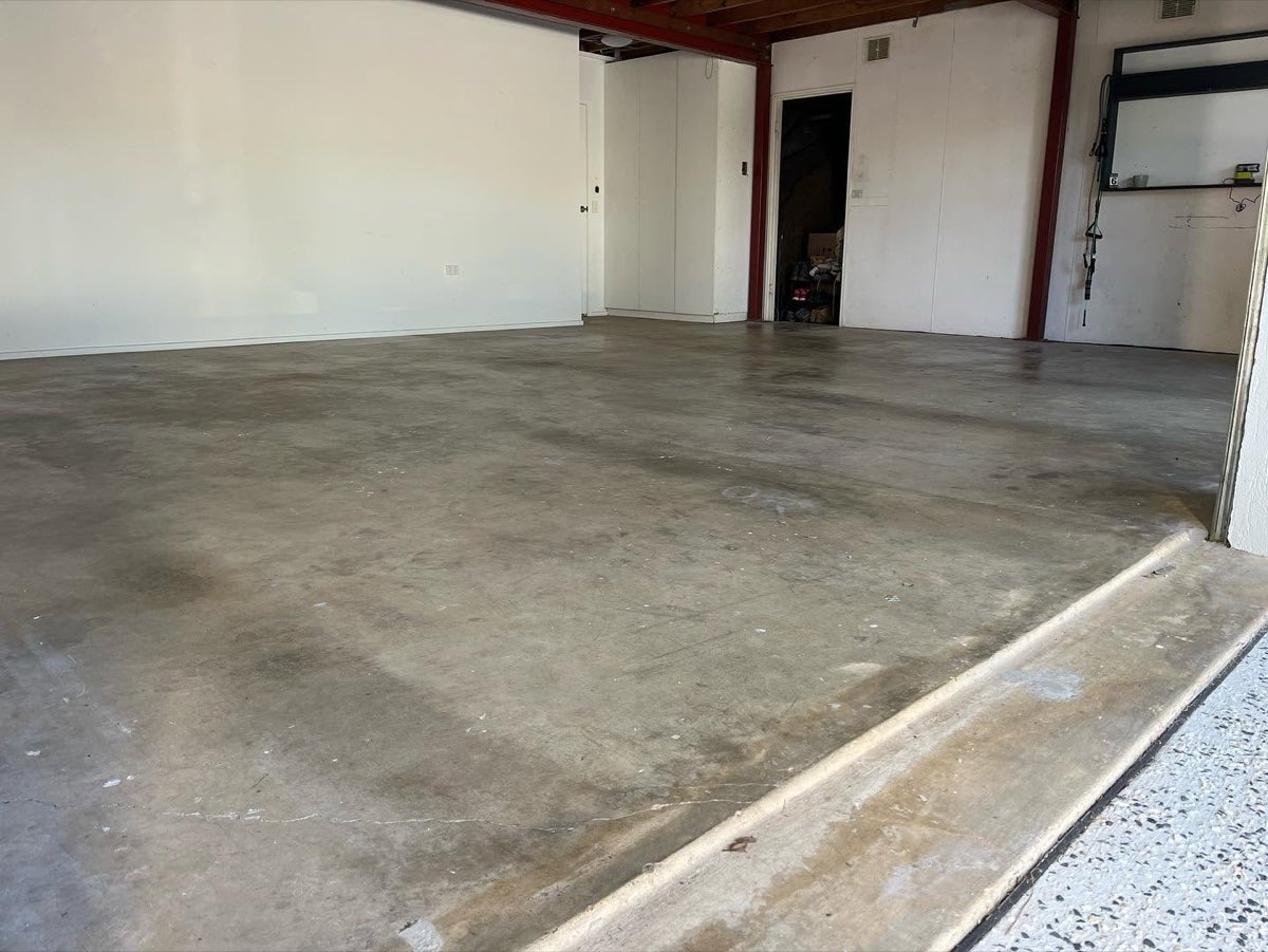 Garage before coating, bare grey concrete marked with old stains, under a timber rafter ceiling with red steel posts and white sheeted walls.