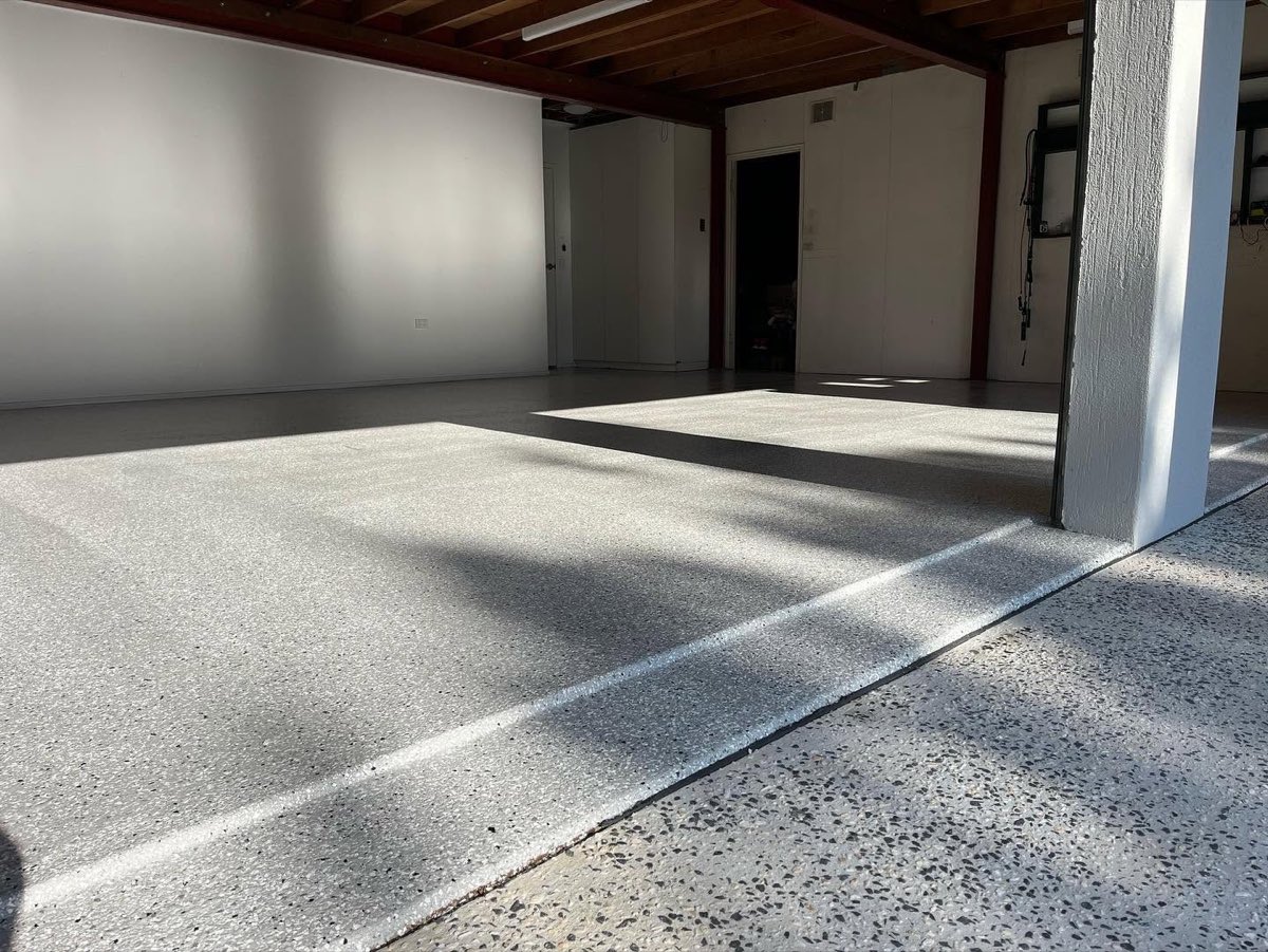 The same garage after coating, a light grey and white flake floor with sunlight falling across it in patches from the open roller door.