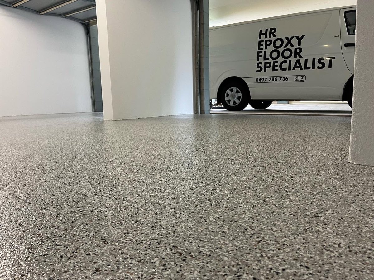 Low view across a finished grey and white flake garage floor, with the HR Epoxy Floor Specialist van parked just outside the open roller door.