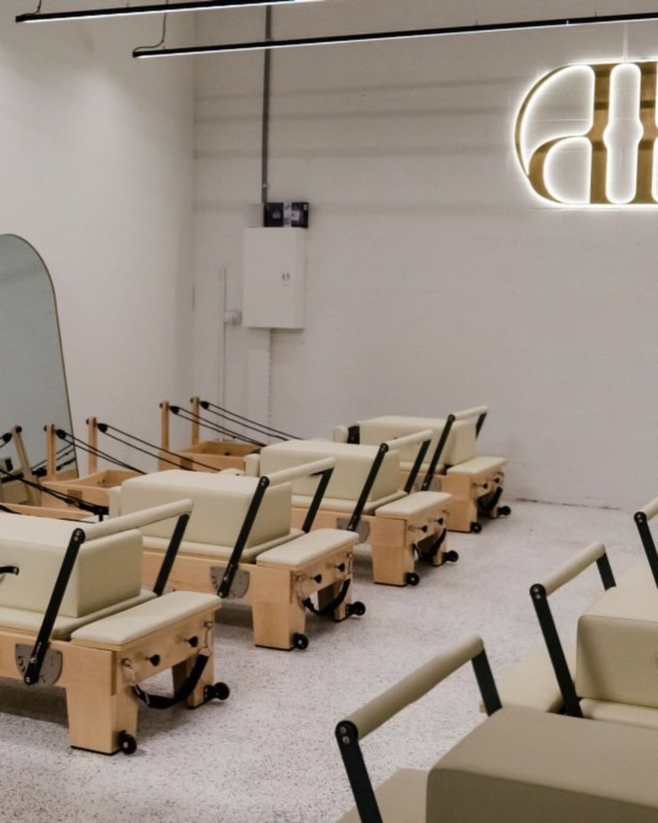 Pilates studio with a cream and white flake floor, rows of timber reformer beds facing a tall arched mirror and a gold illuminated logo on the white wall.