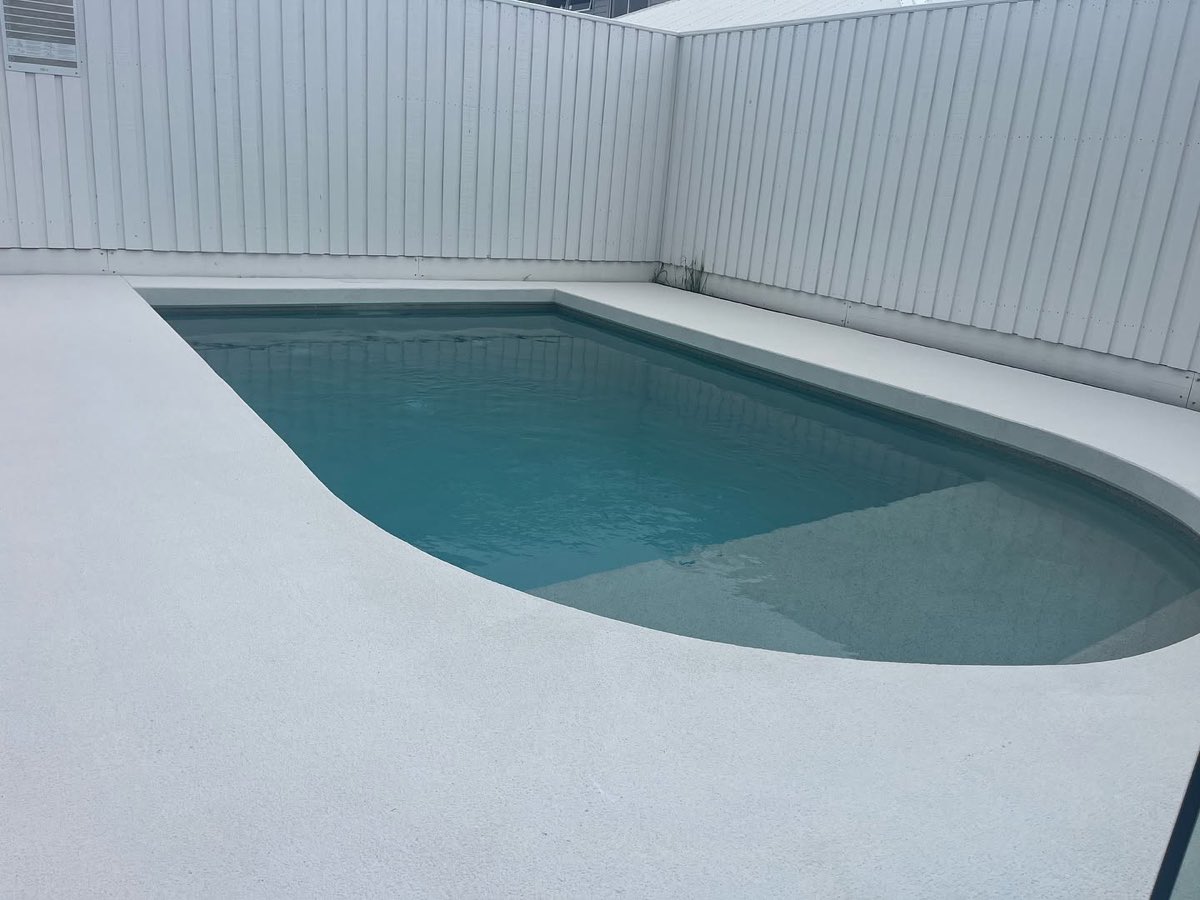 Pool surround coated in a white textured non slip finish, wrapping the curved edge of a pool with teal water, inside a white colorbond fence.