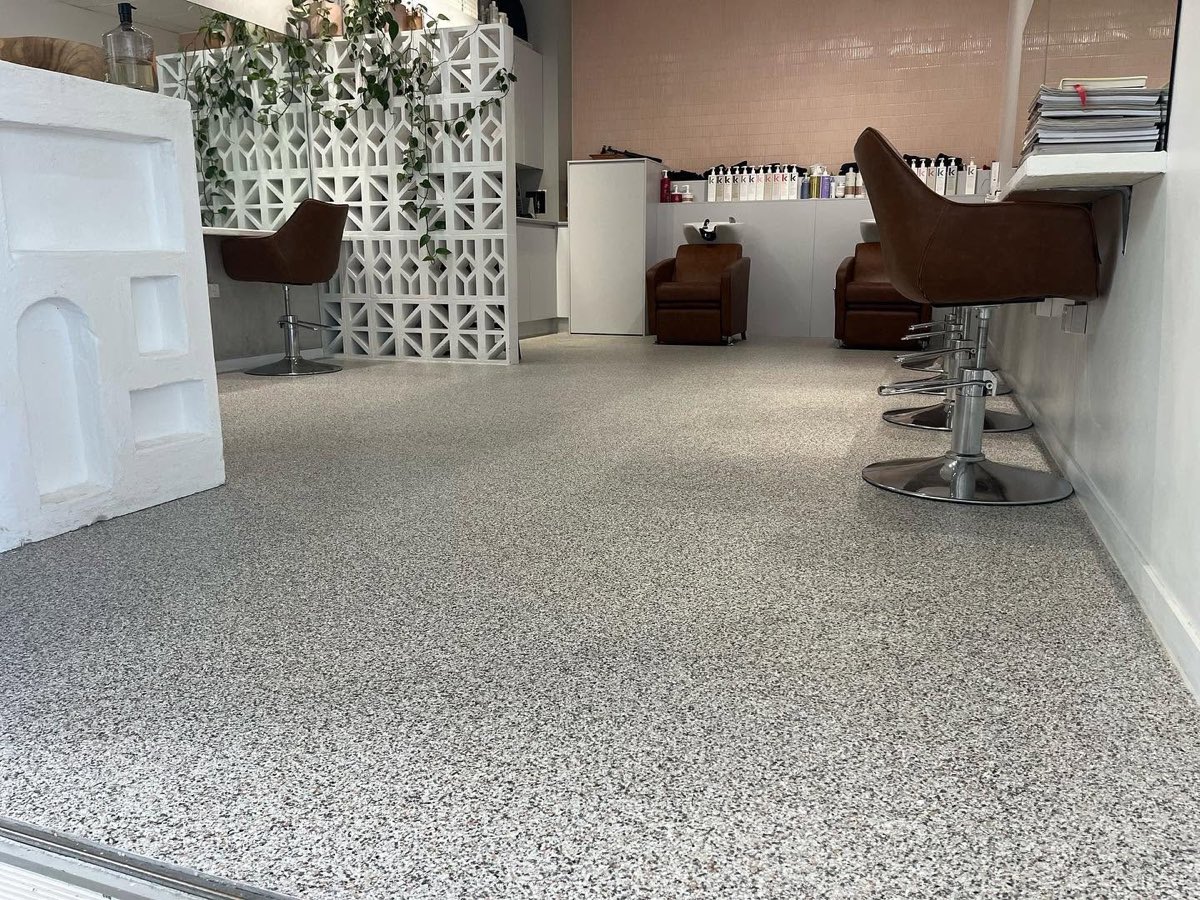 Hair salon interior with a grey and white flake floor running wall to wall, leather styling chairs along the right hand mirror bench and a white breeze block screen at the back.