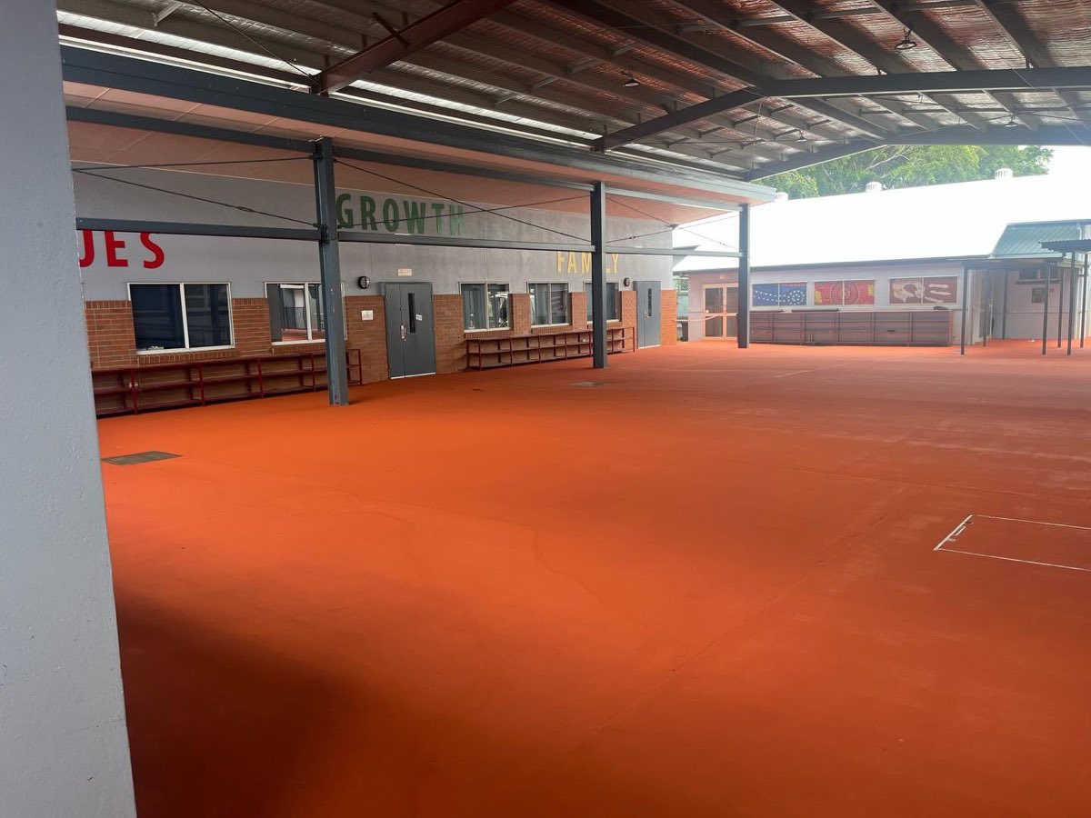 Large covered outdoor school area finished in a bright orange coating, with grey steel posts, brick classroom walls and painted lettering above the windows.