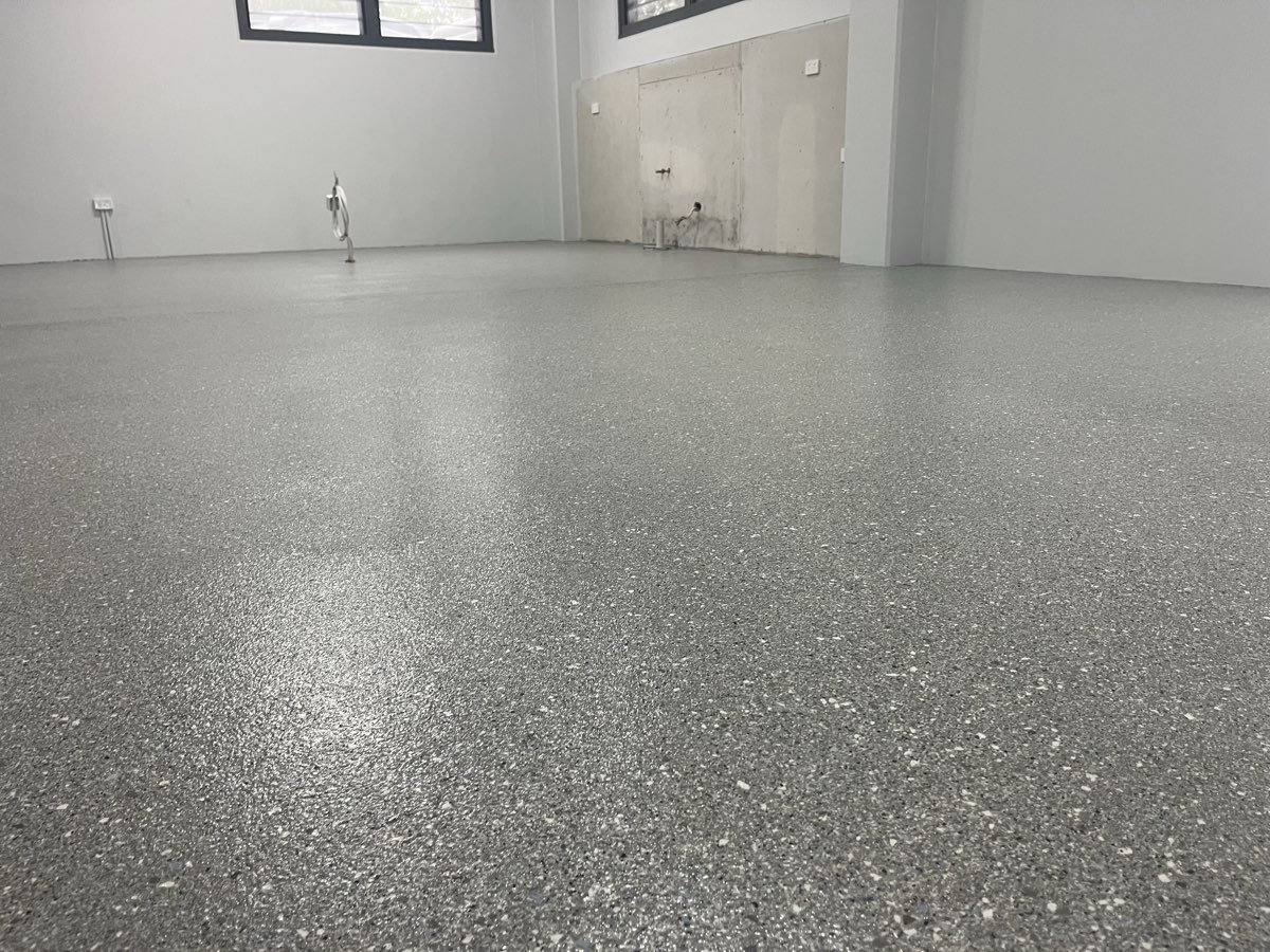 Large empty ground floor room with a glossy grey flake floor reflecting the light, white walls and a sliding door frame at the back.