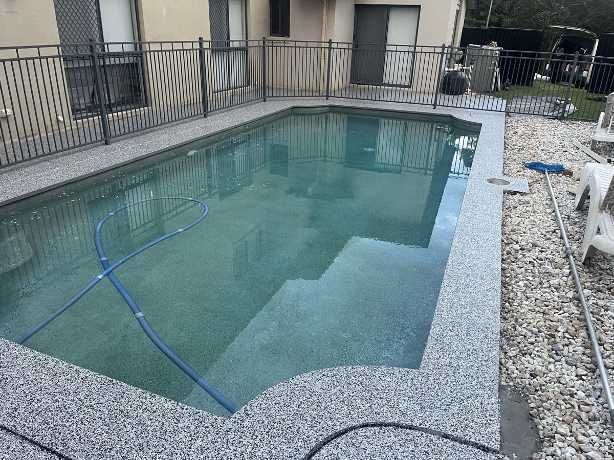 Curved pool edge and surround finished in a grey and white flake coating, with river pebbles and a pool fence alongside.