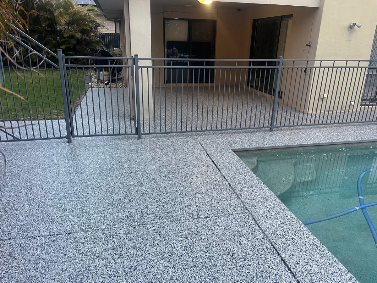 Back patio and pool edge coated in a grey and white flake finish, running up to a black pool fence and gate outside the house.